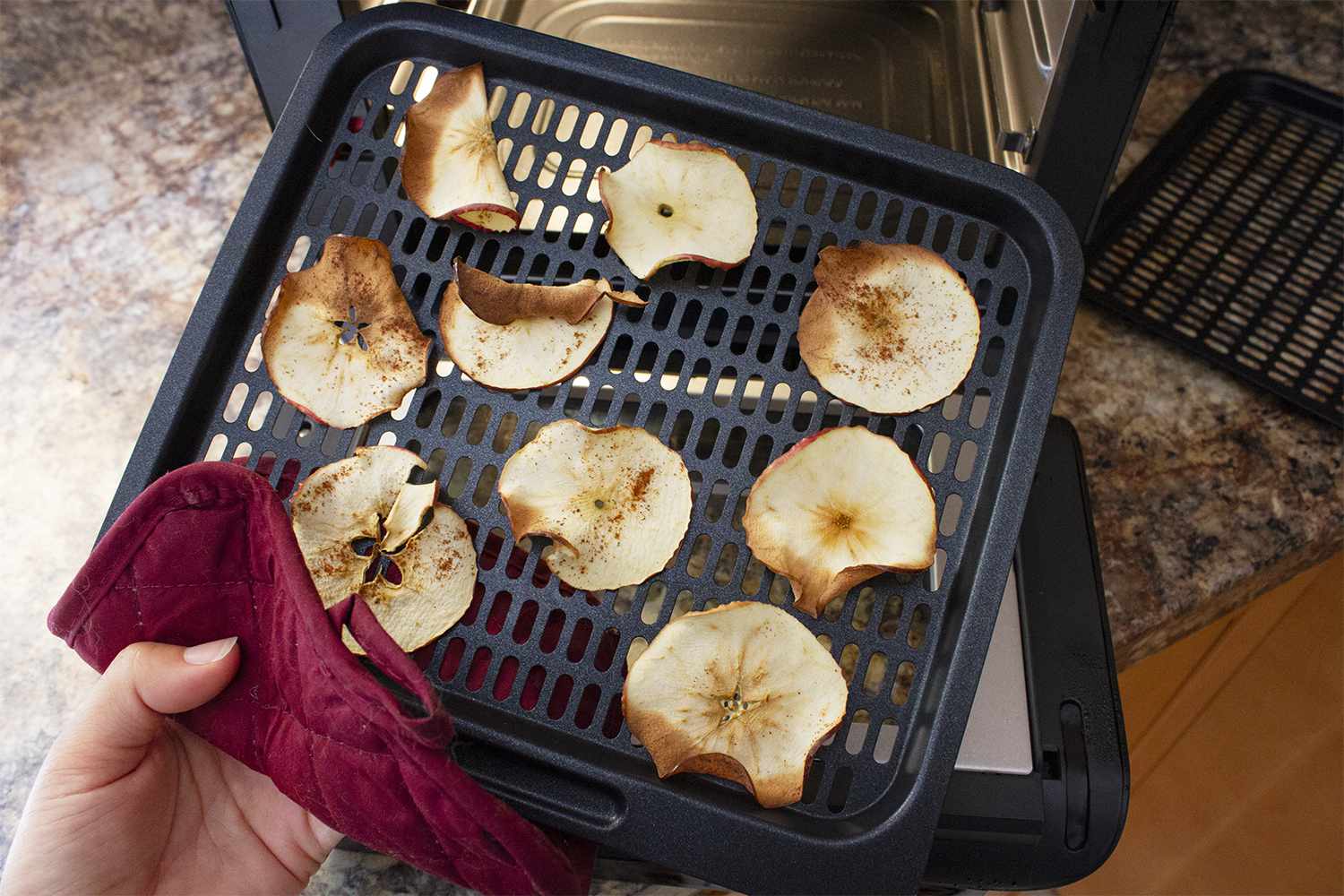 A hand using a towel to hold a tray of baked apple slices in front of the Instant Vortex Plus 7-in-1 Air Fryer Oven