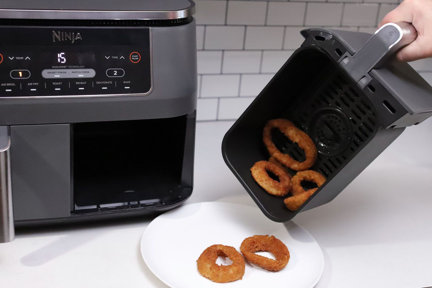 A hand transferring onion rings from the Ninja Foodi 6-in-1 8-Quart 2-Basket Air Fryer on to a white dinner plate
