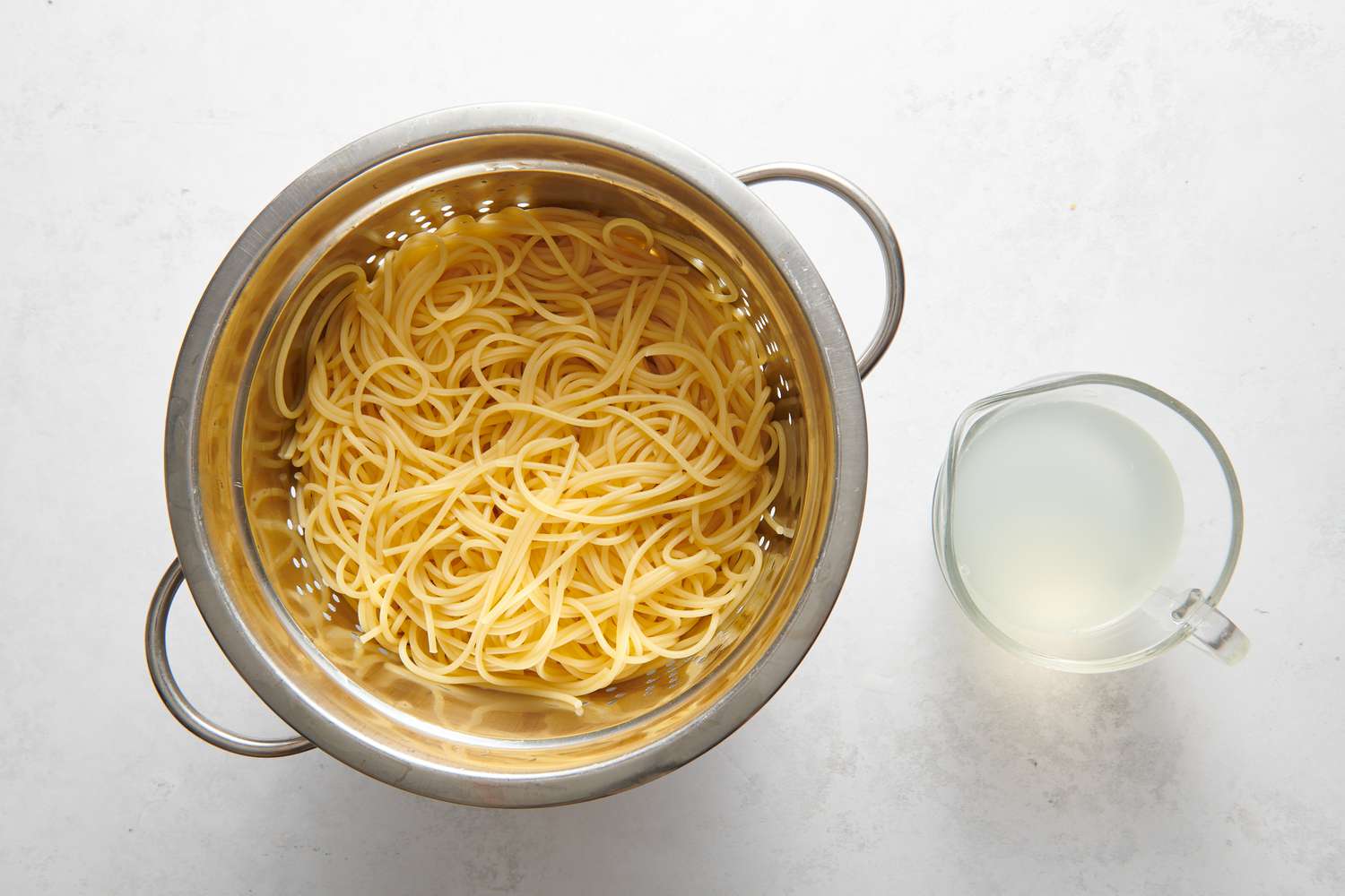 A colander of cooked spaghetti next to a measuring cup of pasta water