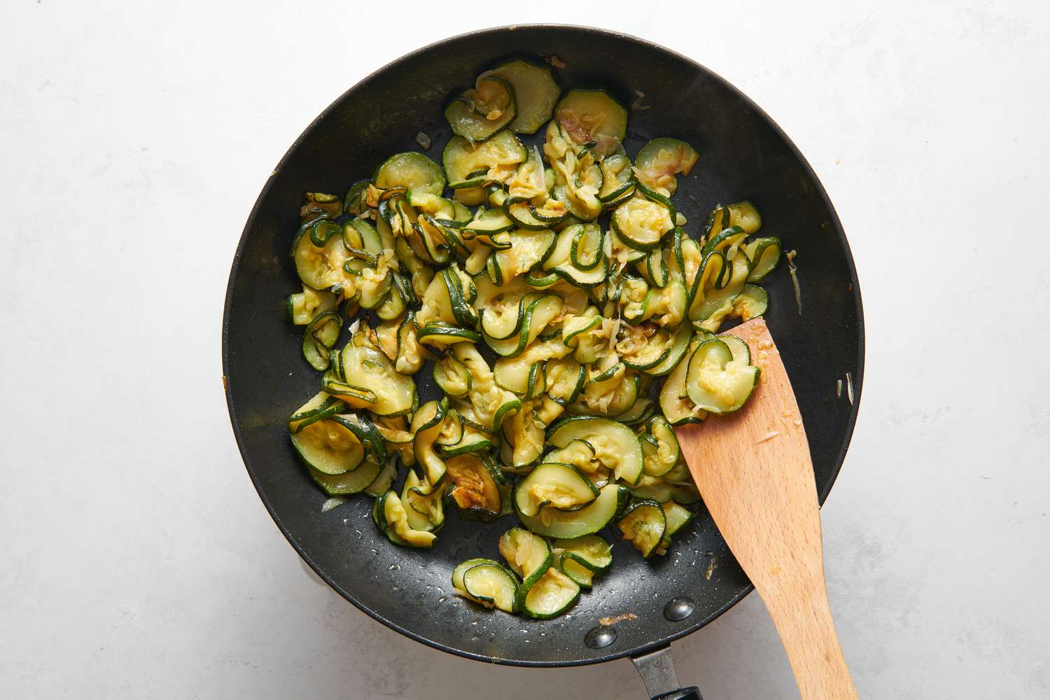 A skillet of sliced zucchini and shallots cooking in olive oil, being stirred with a wooden spatula