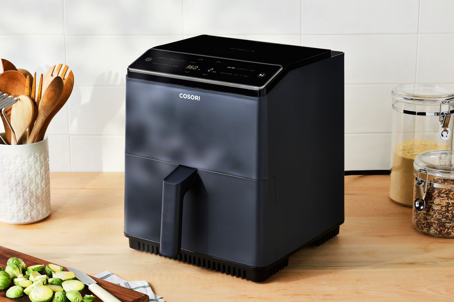 A Cosori Dual Blaze Smart Air Fryer displayed on a wood countertop among cooking ingredients