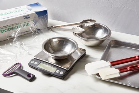 Plastic wrap, Y peeler, digital kitchen scale, metal mixing bowls, metal tongs, rubber spatulas, and a sheet tray