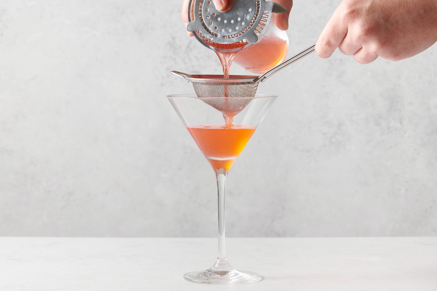 A hand pouring a strawberry martini through two strainers into a martini glass from a cocktail shaker