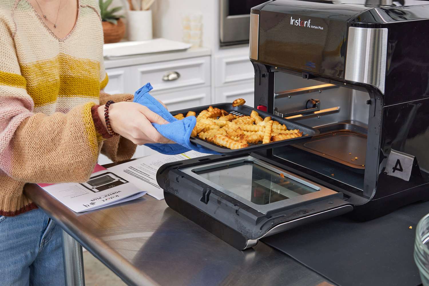 A hand removing a baking sheet full of French fries from the Instant Vortex Plus Air Fryer displayed on a kitchen island