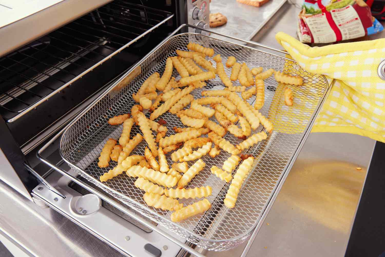 An oven mitt clad hand pulling out a basket of crinkle fries from the Breville Smart Oven Air Fryer