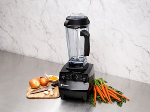 A Vitamix blender on a stainless steel countertop next to a cutting board with onions and garlic cloves on one side and a bunch of carrots on the other side.
